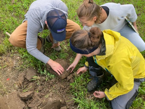 Drei Menschen hocken zusammen auf einem Acker und untersuchen den Erdboden.