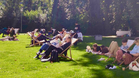 Menschen sitzen in Liegestühlen oder liegen auf Decken auf einer Wiese und lesen.