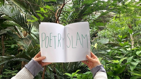 Eine Person hält ein Blatt Papier mit der Aufschrift Poetry Slam vor dem Pflanzendickicht in der Stadtgärtnerei in die Höhe.