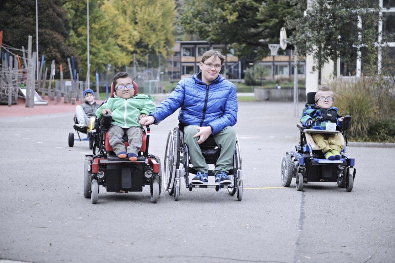 Foto von vier Schülern der SKB. Drie Schüler fahren im Rollstuhl auf dem Pausenplatz. Ein vierter Schüler fährt in einem Dreirad im Hintergrund.