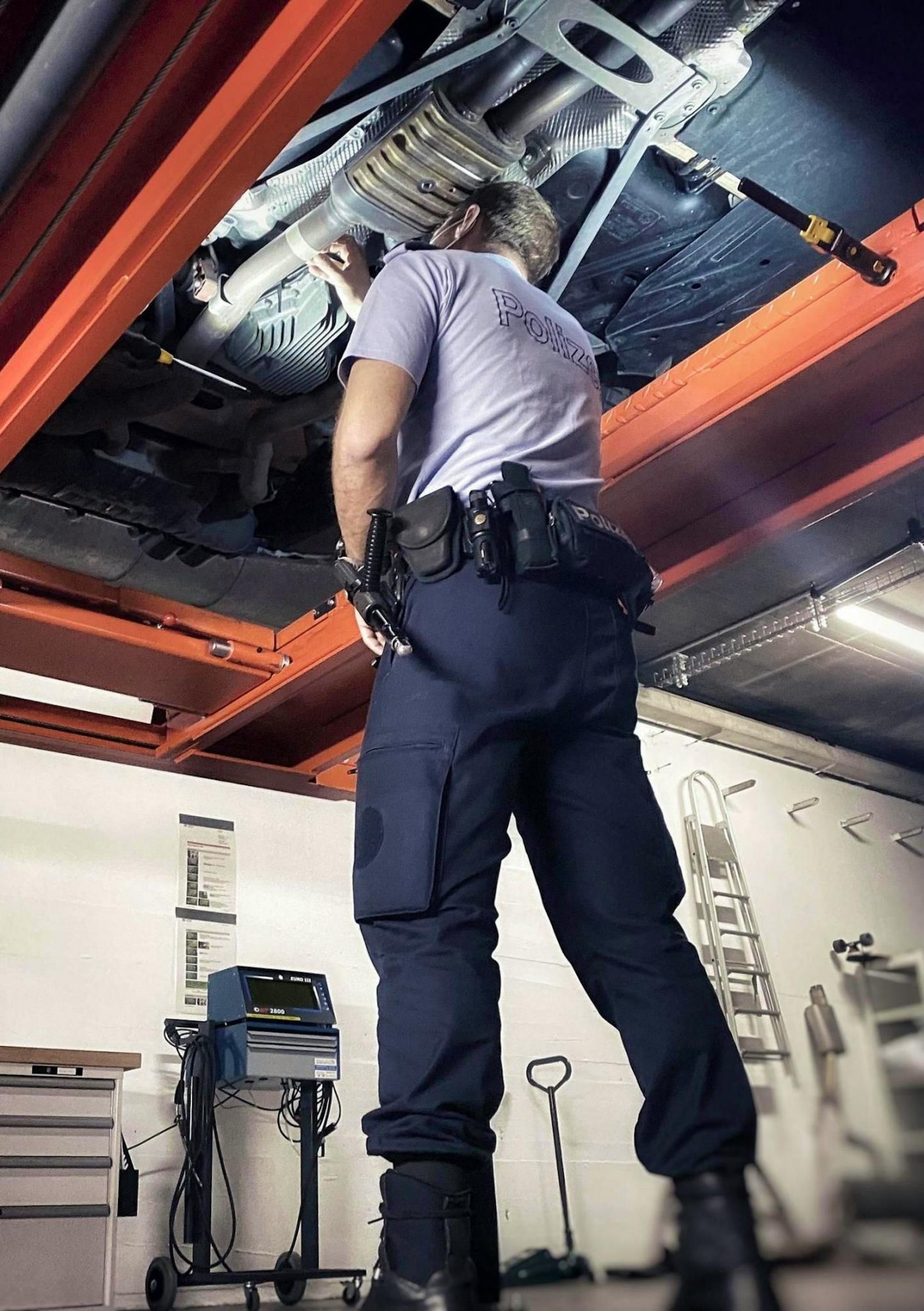 Mitarbeiter der Stadt Polizei Zürich inspiziert die Auspuffanlage eines angehobenen Fahrzeugs in einer Werkstatt.