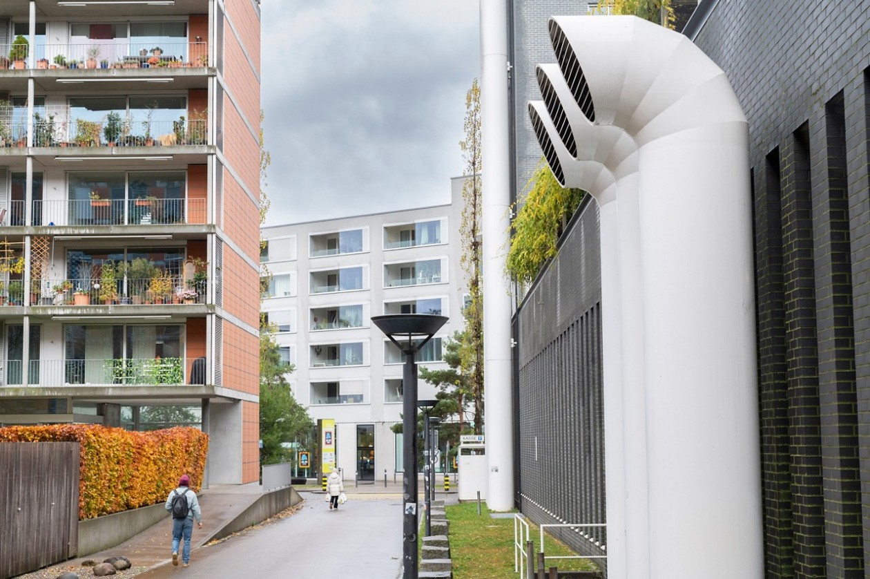Grosse weisse Lüftungsrohre an einem Gebäude neben einem Fussweg in einem Wohngebiet mit mehrstöckigen Wohnhäusern.