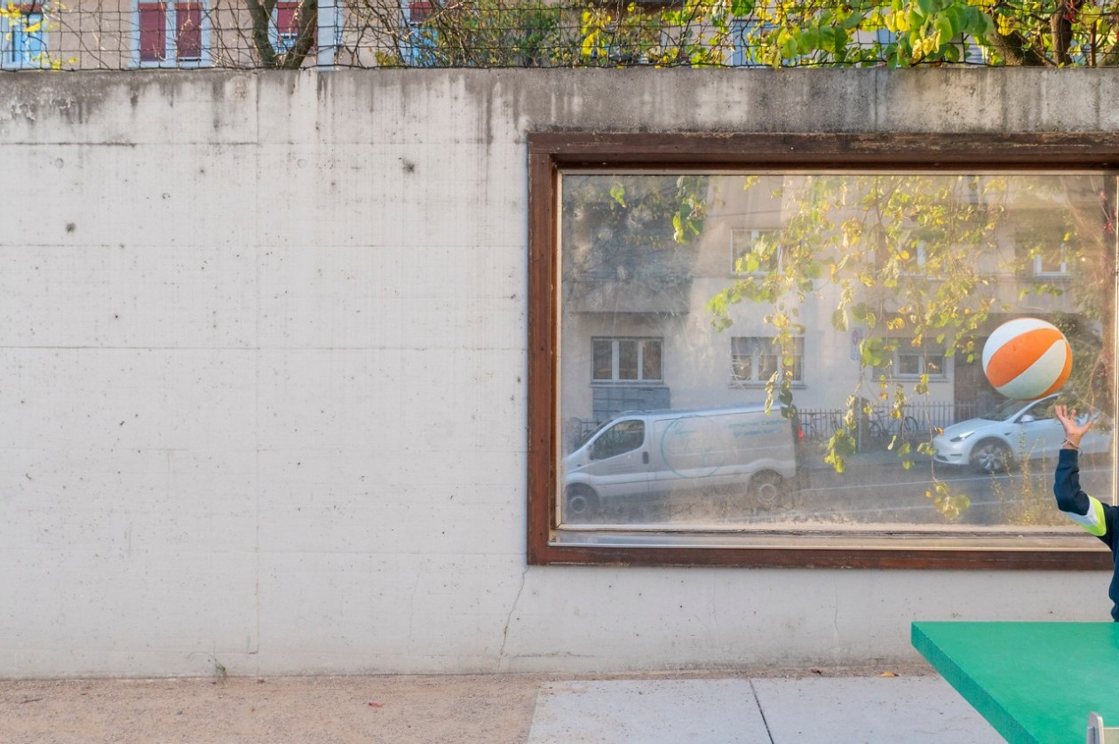 Fenster in einer Mauer eines Schulhofs. Im Vordergrund Pausenspiel von Kindern, im Hintergrund durch das Fenster erblickt man Strassenverkehr.
