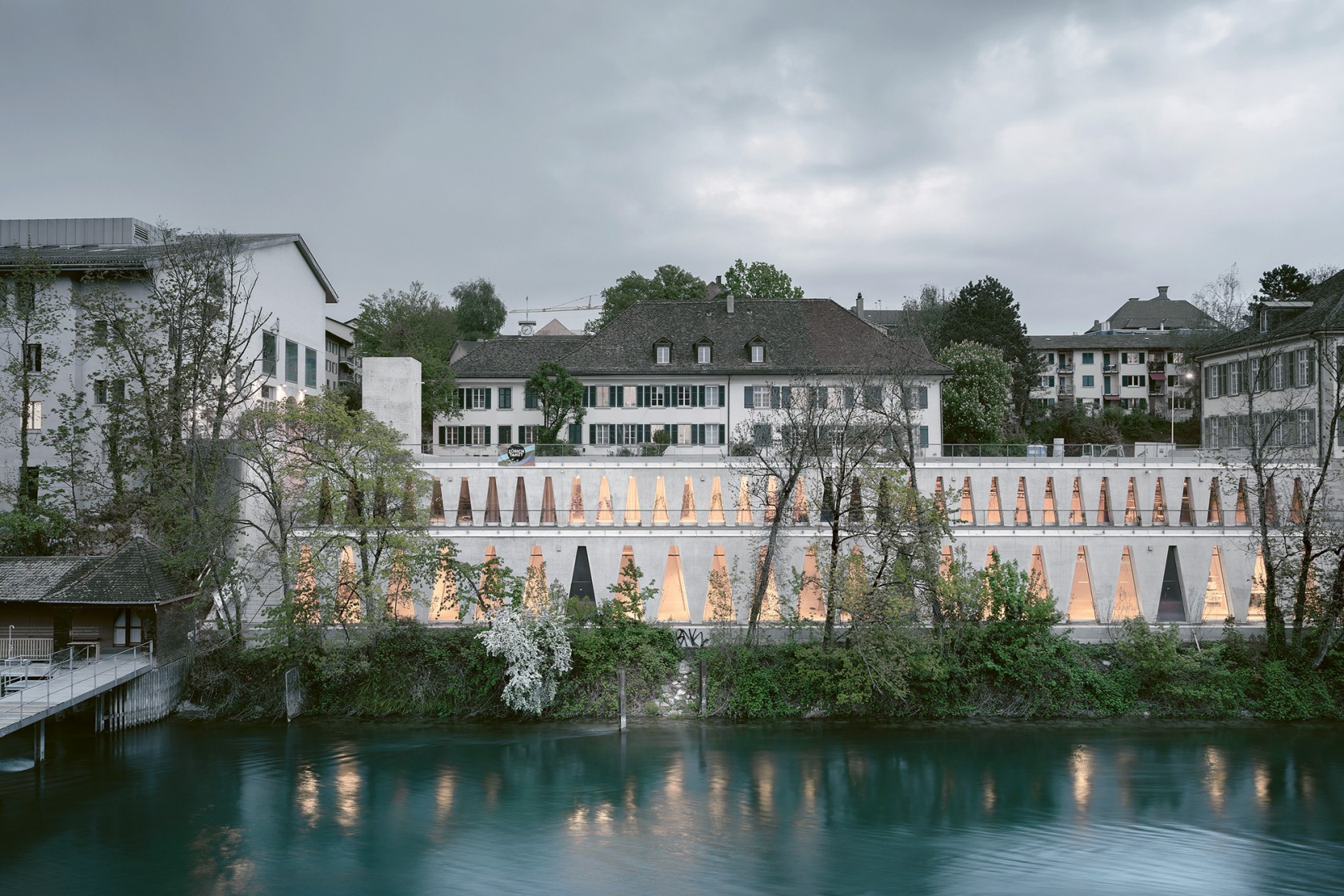 Das Foto wirft einen Blick über die Limmat auf das beleuchtete gut in die Umgebung integrierte Geäude mit den markanten dreieckigen Fenstern.