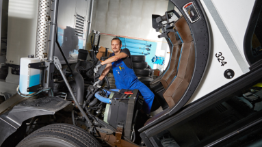 Automobil-Fachmann in blauer Arbeitskleidung auf dem Motor eines Nutzfahrzeugs sitzend