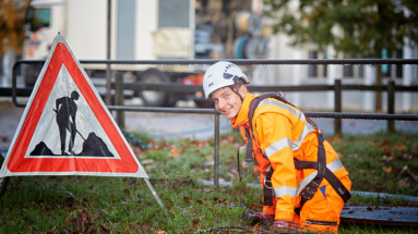 Entwässerungstechnologe in oranger Arbeitskleidung bei der Rohreinigung. Im Hintergrund eine Baustellen-Warntafel.