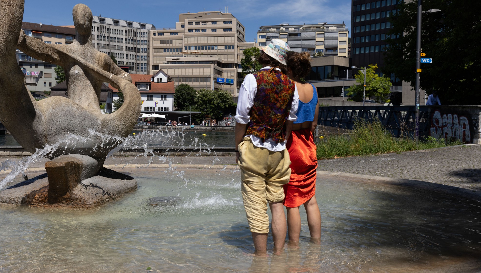 Zwei erwachsene Personen in einem Brunnen stehend mit Hochhäusern im Hintergrund.